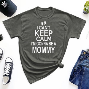 May include: A dark gray t-shirt with the text "I can't keep calm I'm gonna be a mommy" in white lettering with a pair of baby footprints above the text.