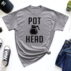May include: A gray t-shirt with a black graphic of a coffee pot and the text "POT HEAD". The shirt is laid flat on a white surface with other items, including a pair of jeans, a hat, sunglasses, and sneakers.
