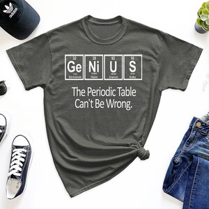 May include: A dark gray t-shirt with a white graphic of the periodic table elements spelling out the word "Genius". The text below the graphic reads "The Periodic Table Can't Be Wrong."