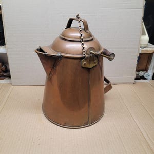 May include: A vintage copper kettle with a lid, handle, and spout. The kettle has a chain connecting the lid to the handle. The copper has a warm, reddish-brown color, and the kettle appears to be well-used.