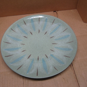 Puede incluir: Un plato redondo de cerámica verde claro con una superficie moteada. El plato presenta un patrón de diseños de rayos de sol estilizados azules y marrones que irradian desde el centro. Un plato de estilo vintage para servir comida.