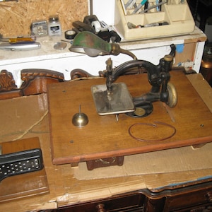Pode incluir: Uma máquina de costura vintage com uma base de madeira e um pedal de metal. A máquina é preta e tem uma lâmpada verde presa a ela. A máquina está sentada em uma caixa de papelão.