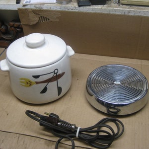 May include: A white ceramic casserole dish with a lid, decorated with a yellow fork and black spoon design. A chrome warming plate and a black power cord are also visible. The set is designed for cooking and serving.