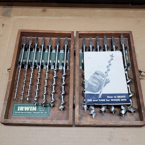 May include: A set of metal auger drill bits arranged in a wooden storage case. The left side features six bits, while the right side contains six additional bits and a booklet titled How to SELECT USE and CARE for WOOD BITS. The Irwin brand logo is visible on a green label inside the case.