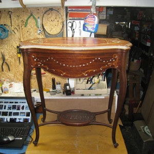 May include: A wooden table with intricate marquetry inlay, featuring a curved top and cabriole legs. The table has a lower shelf and is finished in a rich brown color.