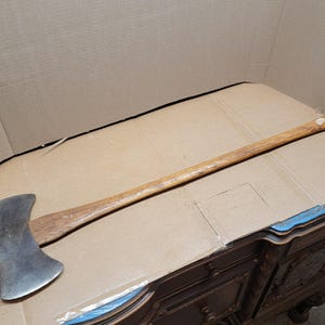 May include: A double-bit axe with a wooden handle and a steel head. The axe head is silver, and the handle is a light brown color. The axe is lying on a brown surface.