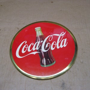 Puede incluir: Letrero redondo de Coca-Cola con fondo rojo y borde dorado. El logotipo de Coca-Cola está en escritura blanca, con una ilustración de botella en el centro.