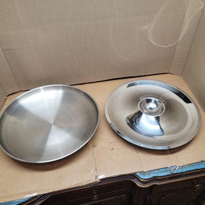 May include: Two stainless steel serving dishes. One is a shallow, round plate with a brushed finish. The other is a domed lid with a central handle. Both pieces are reflective and sit on a cardboard surface.