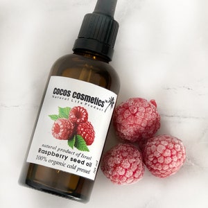 Puede incluir: Una botella de vidrio marrón con un gotero negro y una etiqueta blanca que dice "Cocos cosmetics™ Natural Life Product producto natural de Israel Aceite de semilla de frambuesa 100% orgánico prensado en frío 50 ml". La botella está rodeada de tres frambuesas congeladas.