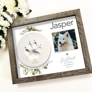 May include: A white wooden frame with a white background. The frame contains a 5 inch paw print, a photo of a white dog, and the text "Jasper", "So Loved", "So Missed", and "2011-2024".