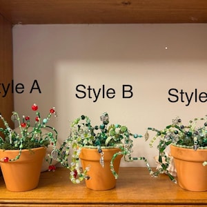 Puede incluir: Tres macetas de terracota con un diseño de planta de cuentas único. El estilo A presenta detalles en rojo y turquesa, el estilo B tiene una disposición verde densa y el estilo C muestra cuentas verdes y blancas.