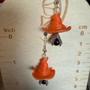 May include: A pair of orange witch hat earrings with a black bead and silver accents. The earrings are suspended from silver hooks. The earrings are approximately 1 inch in length.