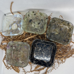 May include: Five square resin dishes with embedded stones and glitter. The dishes are clear, green, and black, with a variety of colors in the stones. The dishes are arranged on brown crinkle paper.