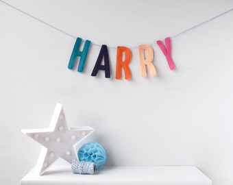 Könnte beinhalten: Ein Filzbanner mit dem Namen "HARRY" in Regenbogenfarben, der an einer weißen Wand hängt. Ein weißes sternförmiges Licht und ein blauer Papierpompon befinden sich auf einem weißen Regal unter dem Banner.