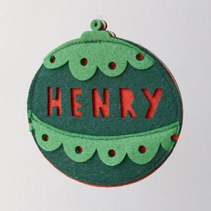 Puede incluir: Adorno navideño de fieltro verde con un recorte de fieltro rojo del nombre "HENRY" en el centro.