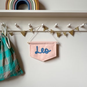Pode incluir: Uma faixa de feltro rosa com o nome "Leo" bordado em fio azul. A faixa está pendurada em uma prateleira branca com uma guirlanda de triângulos dourados com glitter.