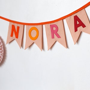 May include: A pink felt banner with the name "NORA" spelled out in orange, red, and yellow felt letters. The banner is strung on a bright orange string.