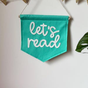 Może przedstawiać: Turkusowy baner z filcu z białym napisem "let's read". Baner jest zawieszony na białej ścianie ze sznurkiem złotych flag z brokatem.
