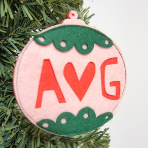 Peut inclure: Ornement de Noël en feutre rose avec une bordure en feutre vert et des points blancs. L'ornement porte les lettres AVG en feutre rouge avec un cœur en feutre rouge au milieu.