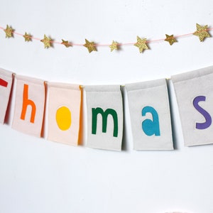 May include: A banner with the name "Thomas" spelled out in colorful felt letters. Each letter is on a separate white flag with a pink border. The flags are strung together with a pink string and are hanging from a white wall. A string of gold glitter stars is strung above the banner.