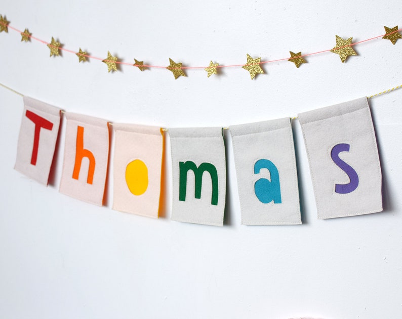 May include: A banner with the name "Thomas" spelled out in colorful felt letters. Each letter is on a separate white flag with a pink border. The flags are strung together with a pink string and are hanging from a white wall. A string of gold glitter stars is strung above the banner.