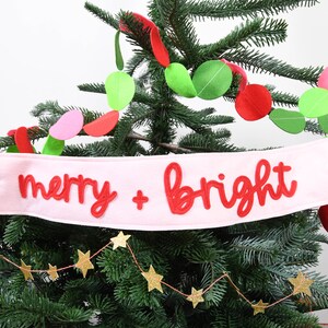 Peut inclure: Un sapin de Noël décoré avec une bannière rose portant l'inscription "merry + bright" en rouge. L'arbre est orné de guirlandes en feutre colorées et d'une guirlande d'étoiles dorées.