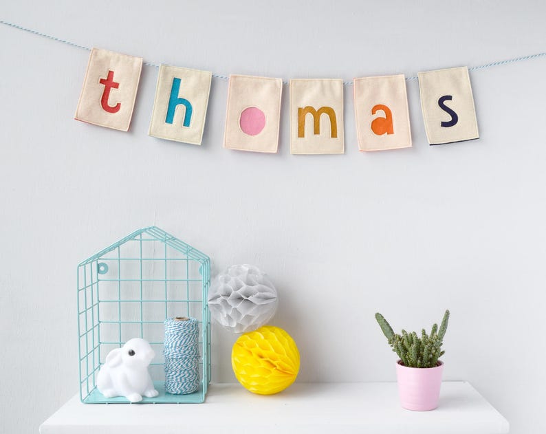 May include: A white fabric banner with the name "thomas" spelled out in colorful felt letters. The banner is hanging on a white wall above a shelf with a blue wire shelf unit, a white rabbit figurine, a ball of twine, a gray honeycomb ball, and a yellow honeycomb ball. A small potted cactus is on the shelf to the right.