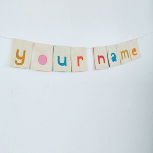 May include: A white fabric banner with the words "your name" spelled out in colorful letters. The letters are cut out of felt and sewn onto the fabric. The banner is strung on a white string.