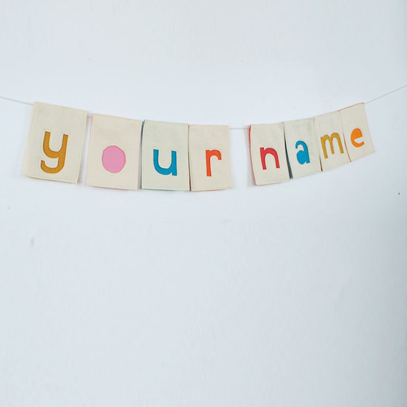 May include: A white fabric banner with the words "your name" spelled out in colorful letters. The letters are cut out of felt and sewn onto the fabric. The banner is strung on a white string.