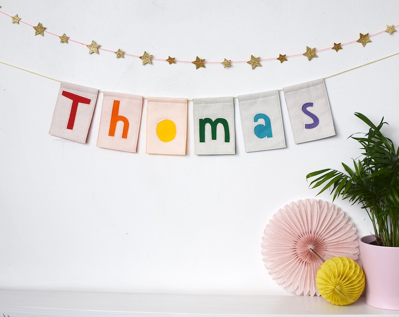 May include: A colorful felt banner with the name "Thomas" spelled out in rainbow letters. The banner is strung with a gold star garland.