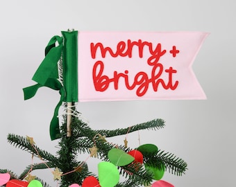 May include: A pink felt banner with the words "merry + bright" in red felt letters, attached to the top of a small artificial Christmas tree.