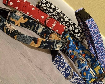 May include: Five different dog collars with various patterns. The collars are made of fabric and have metal buckles. The patterns include red with white polka dots, black with white paw prints, blue with white circles, blue with a jungle animal print, and purple with a swirl pattern.