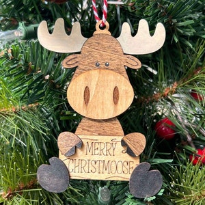 May include: Wooden Christmas bauble shaped like a moose holding a sign that says "Merry Christmoose". The ornament is brown with black hooves and antlers. It is hanging on a green Christmas tree.