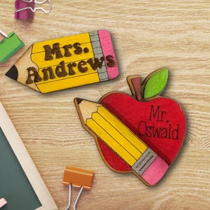 Peut inclure: Deux étiquettes de nom en bois sur le thème des enseignants. L'une est en forme de crayon jaune avec "Mrs. Andrews" en lettres marron. L'autre est en forme de pomme rouge avec un crayon jaune et "Mr. Oswald" en lettres marron.