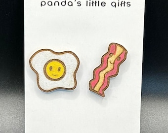 May include: Wooden breakfast-themed earrings featuring a fried egg and bacon. The egg has a yellow yolk and a smiling face. The bacon is pink and tan with a smiling face. The earrings are on a white card with the text "panda's little gifts".