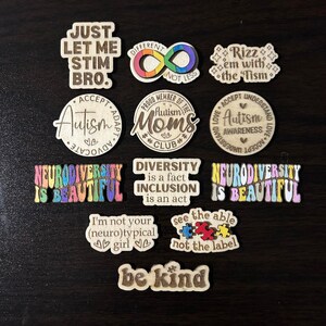 May include: A collection of 12 wooden laser-cut ornaments with various messages about autism, neurodiversity, and inclusion. The ornaments feature phrases like "Just Let Me Stim Bro", "Neurodiversity is Beautiful", "Autism Moms Club", "See the Able Not the Label", and "Be Kind".