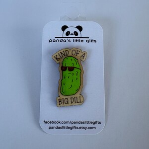 May include: A wooden pin featuring a green pickle wearing sunglasses, with the text "KIND OF A BIG DILL". The pin is attached to a white card with a panda logo and the text "panda's little gifts".