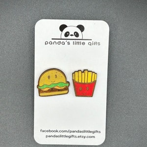 Peut inclure: Un ensemble de deux aimants en bois sur le thème de la nourriture. Le premier aimant est un hamburger jaune et brun avec de la laitue verte et une tomate rouge. Le deuxième aimant est une boîte rouge et jaune de frites. Les aimants sont sur une carte blanche avec le texte "panda's little gifts" et l'adresse du site web "facebook.com/pandaslittlegifts" et "pandaslittlegifts.etsy.com".