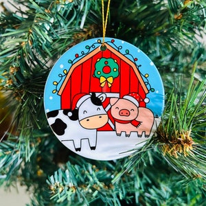 Peut inclure: Une boule de Noël ronde avec une grange rouge, une vache et un cochon devant. La grange a une couronne sur la porte et les animaux portent des chapeaux de Père Noël.