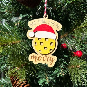 May include: A wooden Christmas ornament shaped like a yellow pickleball with a Santa hat on top. The ornament has the text "Eat, Dink, and Be Merry" written on it.