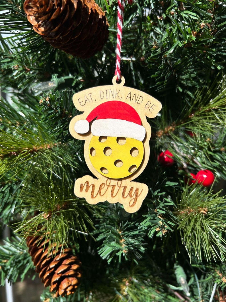 May include: A wooden Christmas ornament shaped like a yellow pickleball with a Santa hat on top. The ornament has the text "Eat, Dink, and Be Merry" written on it.