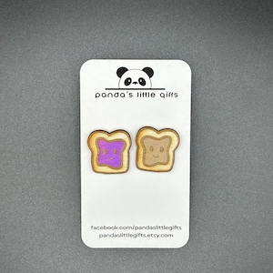 Può includere: Un paio di orecchini in legno a forma di fette di pane con un viso sorridente. Una fetta ha la marmellata viola in cima e l'altra ha il burro di arachidi marrone. Gli orecchini sono su una carta bianca con il testo "panda's little gifts" e l'indirizzo del sito web "pandaslittlegifts.etsy.com".