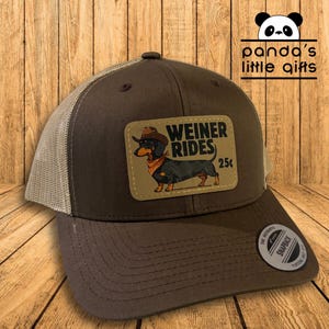 Puede incluir: Gorra de camionero marrón con la parte trasera de malla beige y un parche con un perro salchicha con sombrero de vaquero y pañuelo. El parche dice "WEINER RIDES 25¢". La gorra tiene cierre snapback.