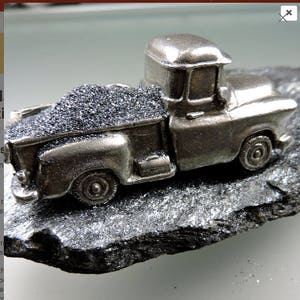 May include: A small, silver-colored pickup truck model with a detailed design. The truck bed is filled with a dark, granular substance. The model sits on a dark, textured surface, creating a contrast between the shiny truck and the rough base.