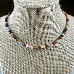 May include: A colorful beaded necklace with a variety of stones, including red, green, blue, and yellow. The necklace is on a fabric-covered mannequin.