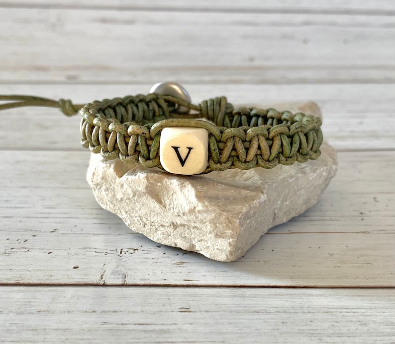 K&ouml;nnte beinhalten: Ein gr&uuml;nes geflochtenes Lederarmband mit einer wei&szlig;en Perle, die den Buchstaben "V" in Schwarz tr&auml;gt.