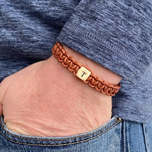 May include: A brown leather braided bracelet with a gold square charm engraved with the letter "T".