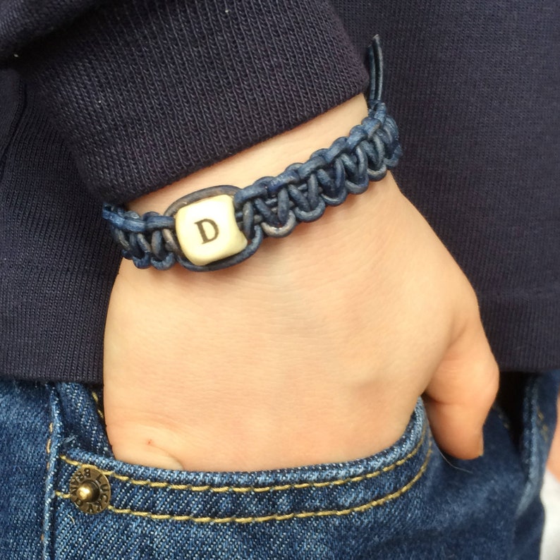 K&ouml;nnte beinhalten: Ein geflochtenes Lederarmband in Marineblau mit einem goldenen Anh&auml;nger mit dem Buchstaben "D".