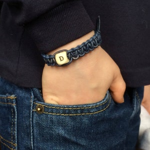 K&ouml;nnte beinhalten: Ein blaues geflochtenes Armband mit einem goldenen Anh&auml;nger, auf dem der Buchstabe 'D' eingraviert ist.