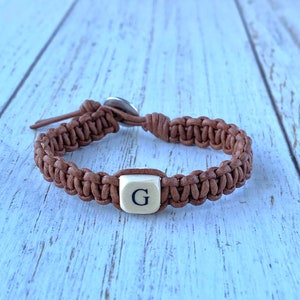 K&ouml;nnte beinhalten: Ein braunes Lederarmband mit geflochtenem Design und einer wei&szlig;en quadratischen Perle mit dem Buchstaben "G" darauf.